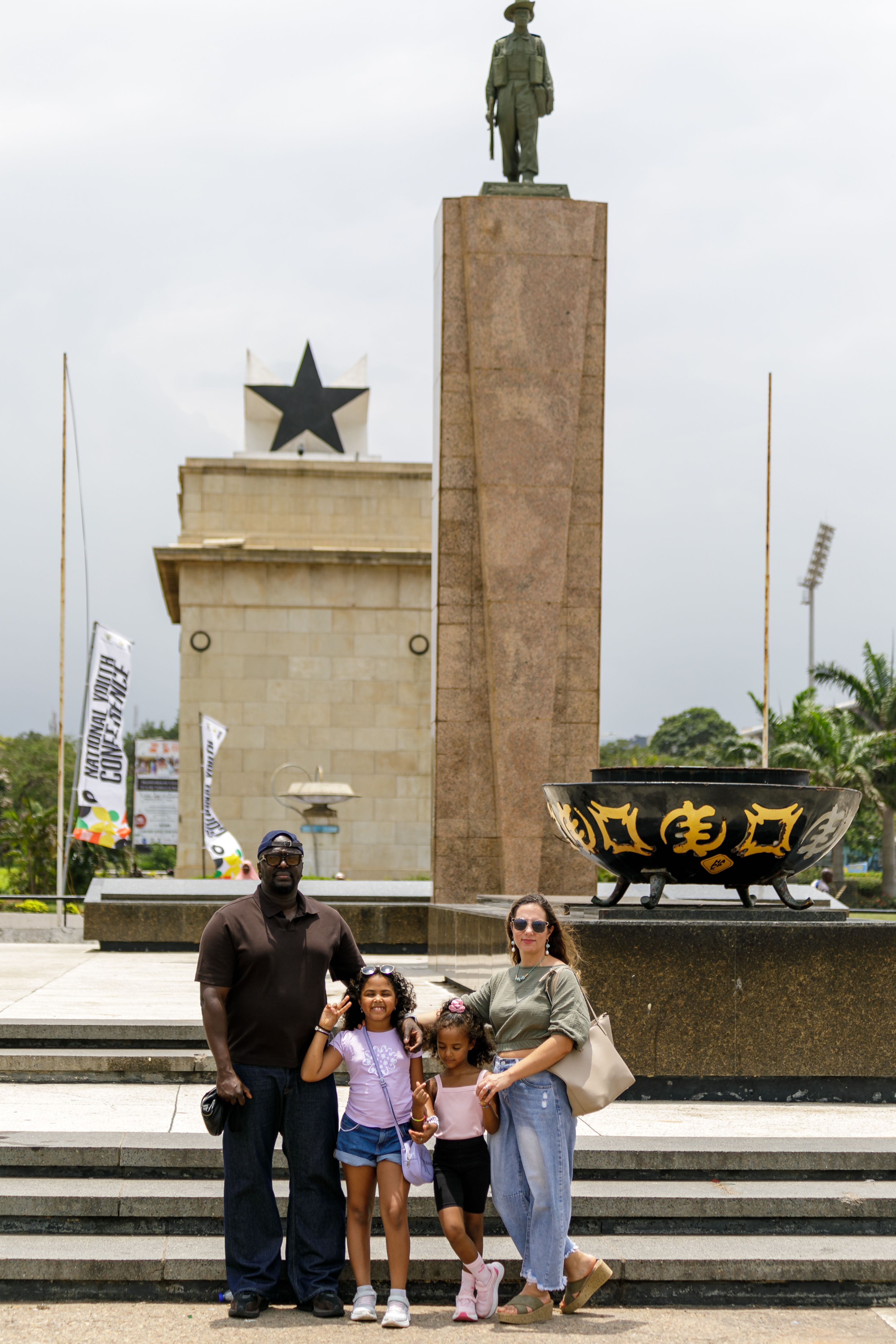 A diverse family posing at the Black Star Square in Accra, featuring iconic landmarks.