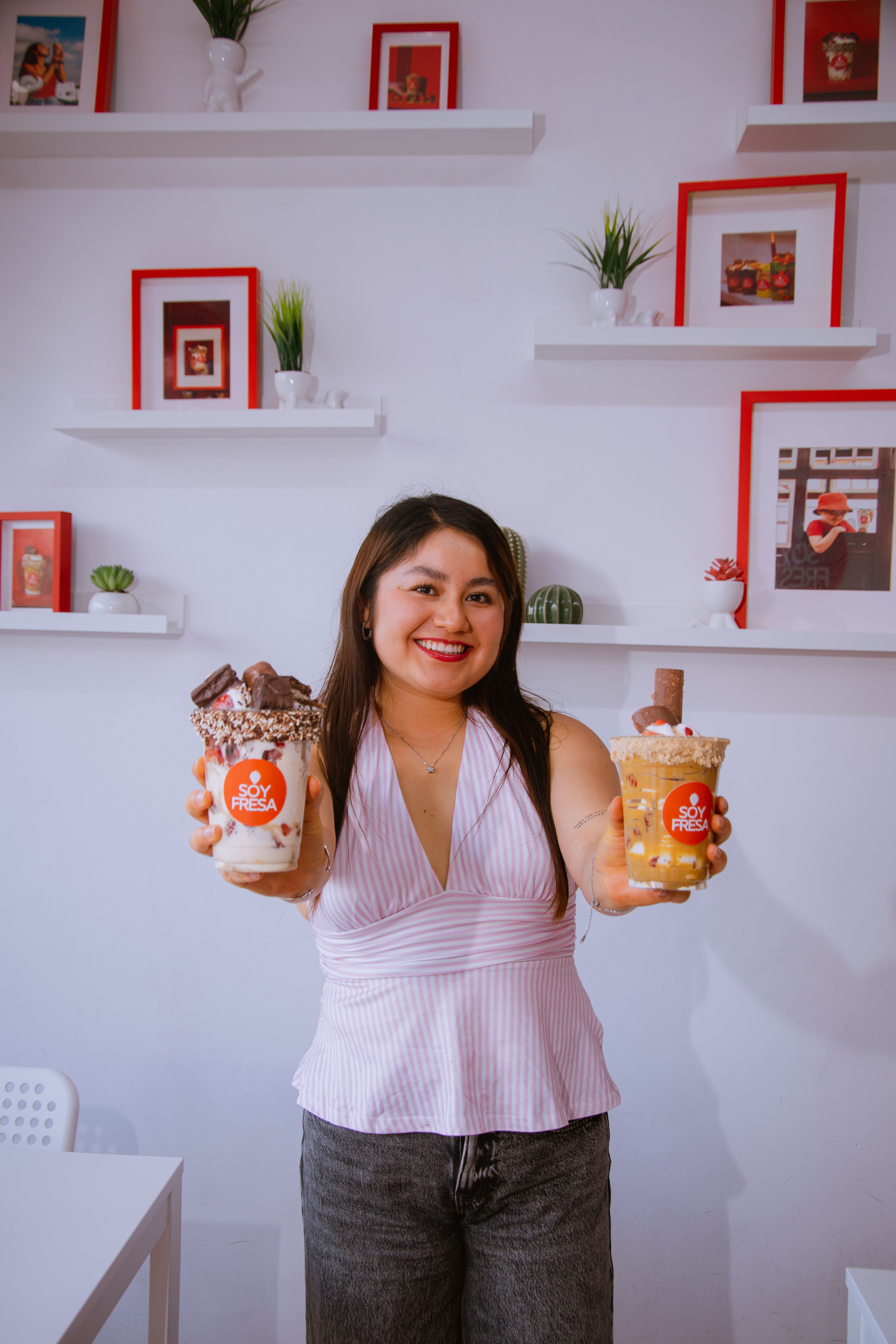 Smiling woman displaying two creamy dessert beverages in a cozy cafe setting with framed decor.
