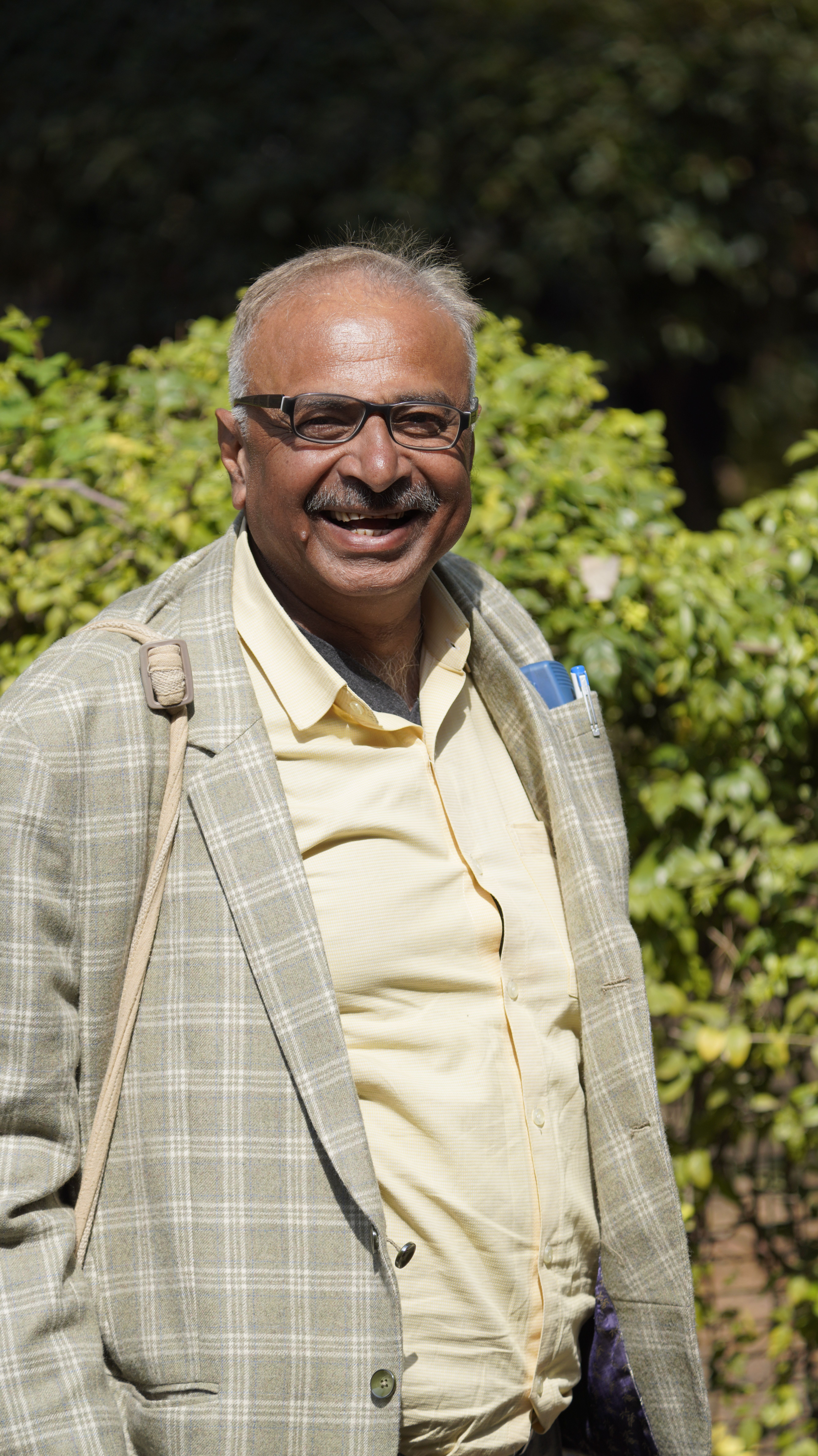 A man in a plaid jacket smiling warmly outdoors, surrounded by greenery.