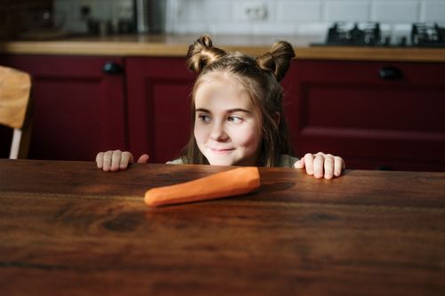 Free A young girl with a playful hairstyle peeks over a wooden table at a carrot in a sunlit kitchen. Stock Photo