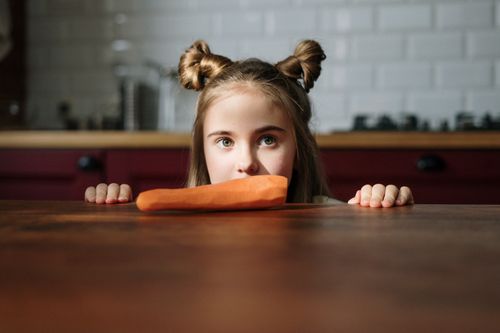 Free Cute girl with hair buns peeking over a table to look at an organic carrot indoors. Happy and playful scene. Stock Photo