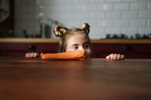 Free A curious child with a cute hairstyle peeks over a kitchen table at a fresh carrot. Stock Photo