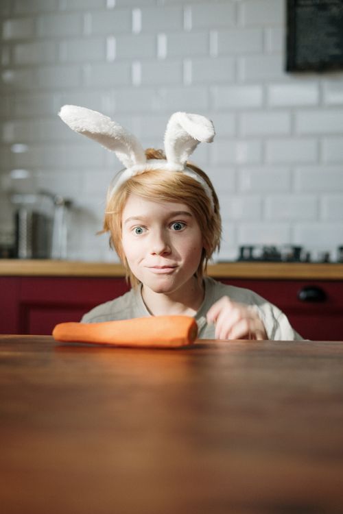 Free A playful child wearing bunny ears looks at a carrot on a kitchen table, capturing a fun indoor moment. Stock Photo