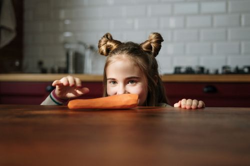 Free A young girl playfully peeks over a kitchen table at a carrot, embodying whimsy and fun. Stock Photo
