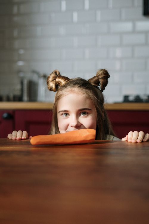 Free Adorable girl with hair buns peeks over a table holding a carrot, indoors. Stock Photo