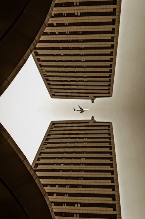 Free Low Angle Photography of Airplane Flying over Brown Concrete Building Stock Photo