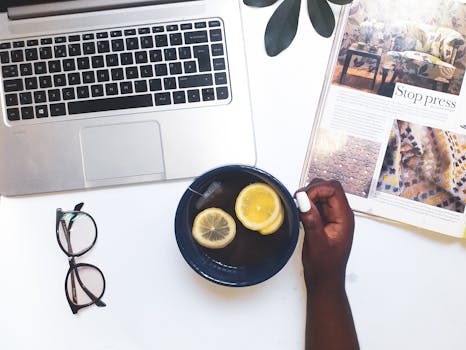 Top view of a modern workspace featuring a laptop, tea with lemon slices, glasses, and a magazine on a white desk.
