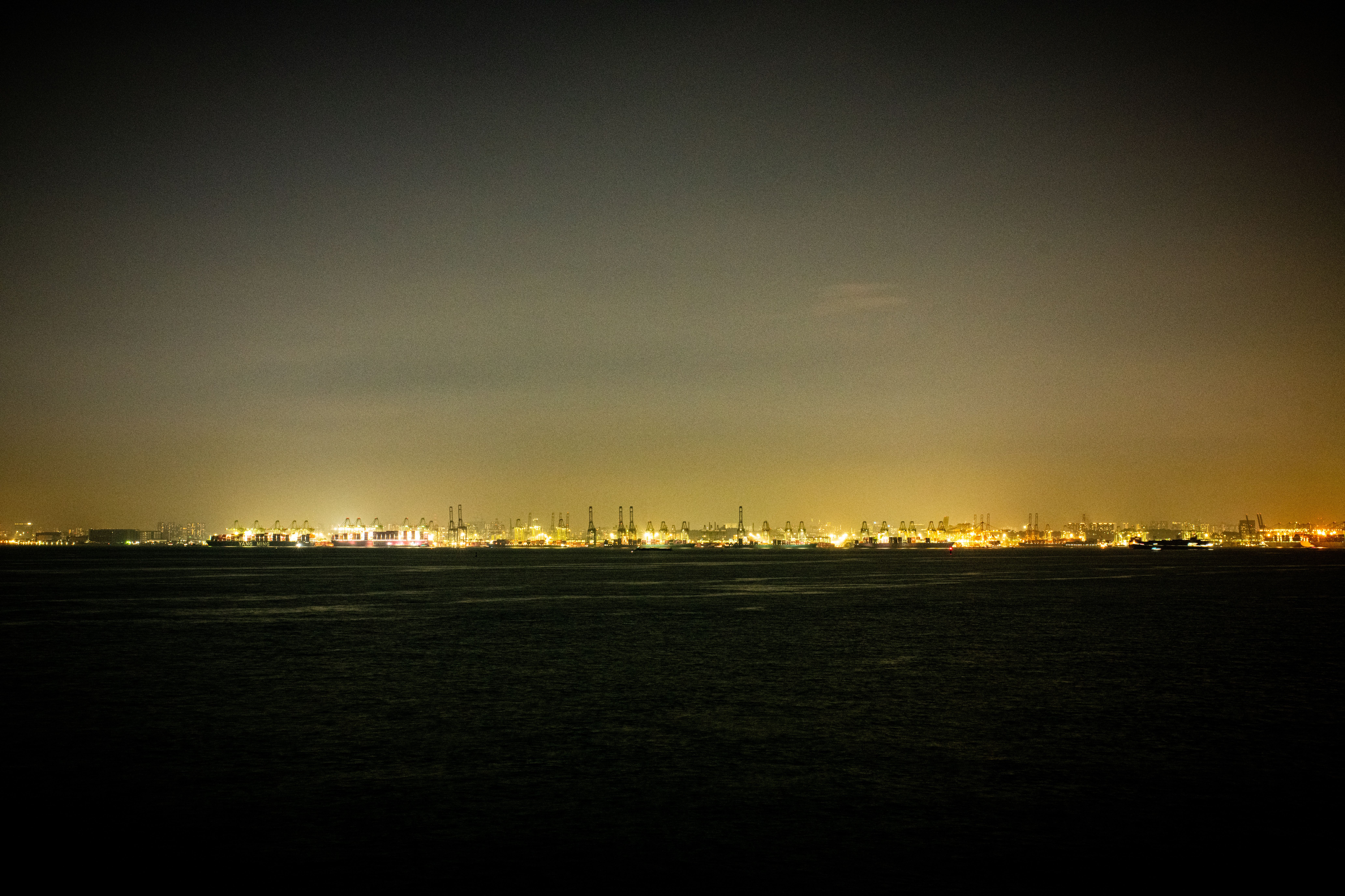 Fotomural Vista nocturna de larga exposición de un paisaje urbano portuario brillantemente iluminado, que muestra estructuras industriales y luces de la ciudad a lo lejos.