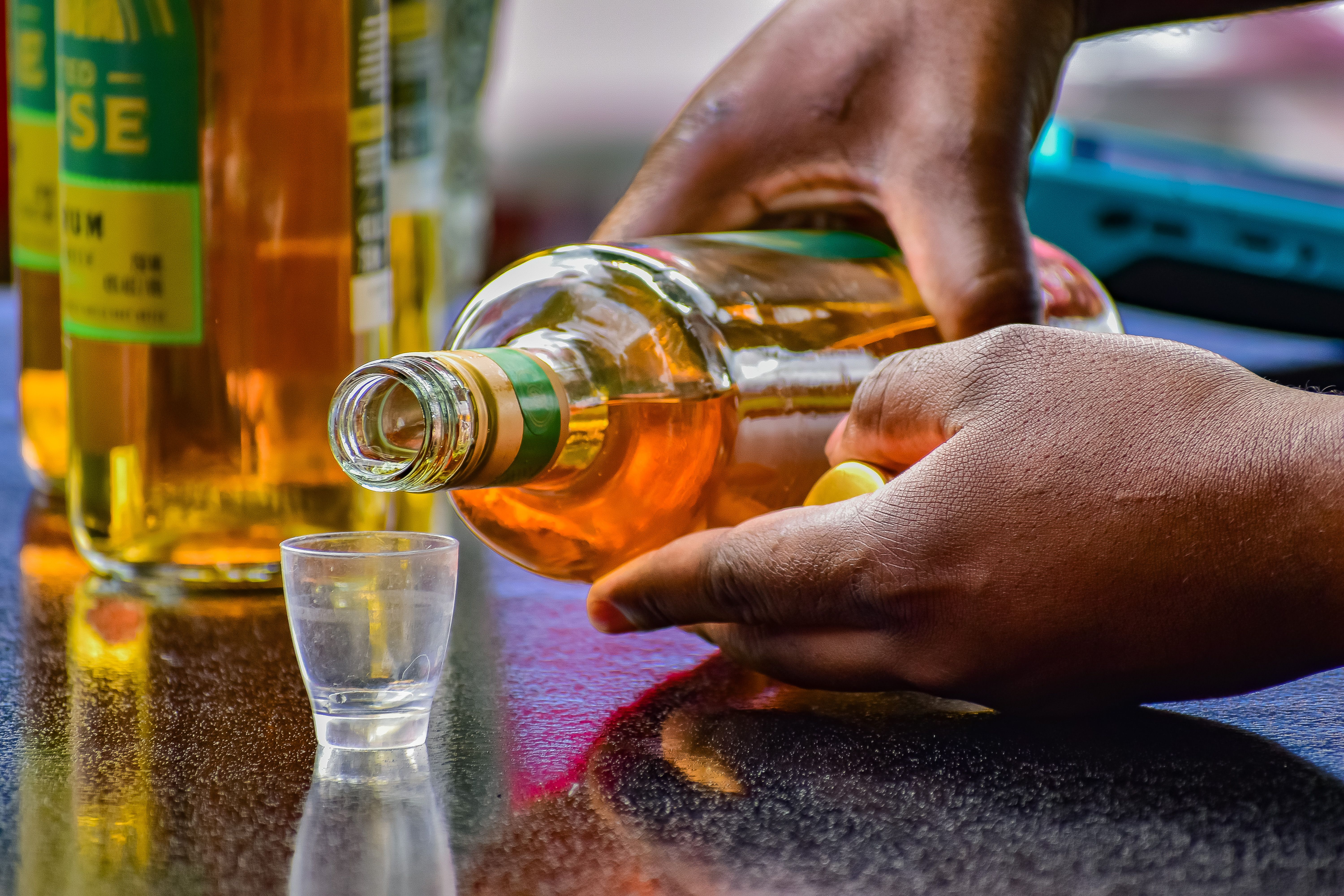 Close-up of hands pouring golden rum into a shot glass, reflecting a casual outdoor setting.