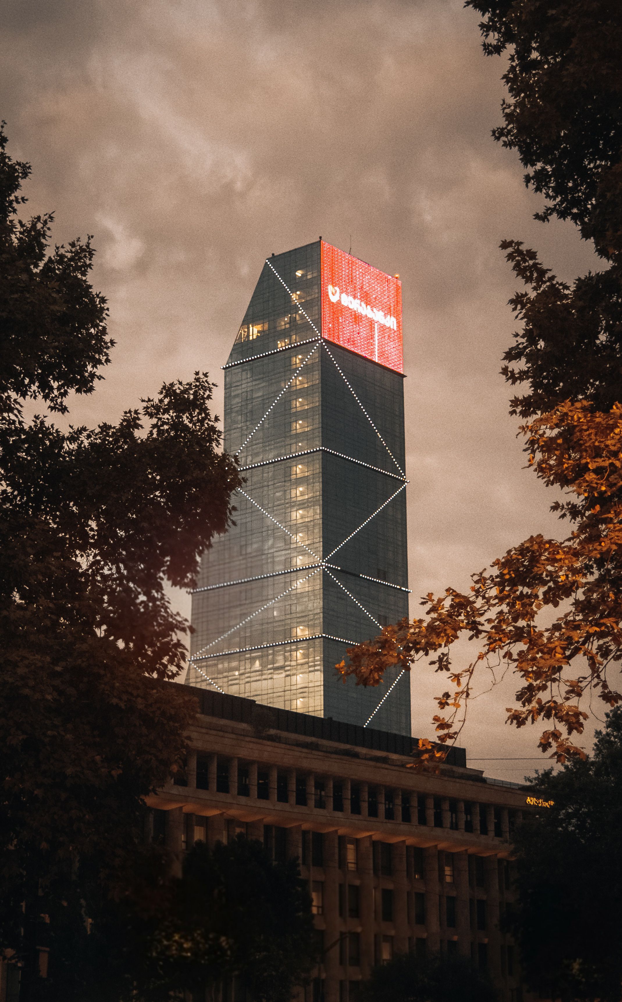 Skyscraper with glowing signage during twilight framed by trees.