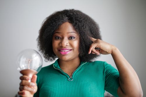 Free Smiling young African American lady in green casual clothes holding light bulb in hand and touching temple while having brilliant idea and looking at camera on gray background Stock Photo
