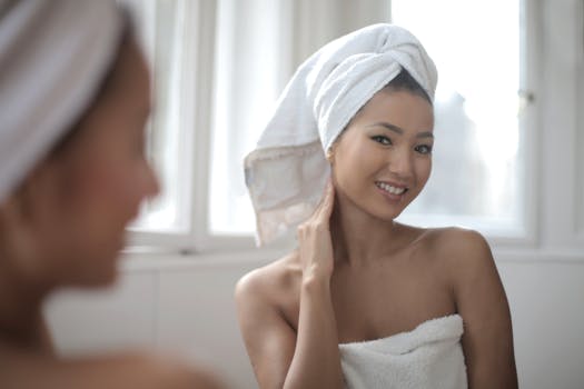 A woman wrapped in a towel smiles at her reflection in a bright bathroom.