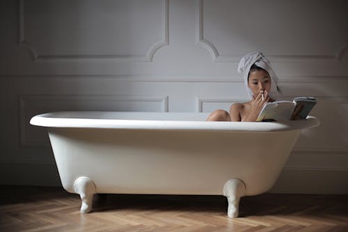 Sensual young Asian lady with towel on head relaxing in bath with magazine while smoking cigarette in modern bathroom at home