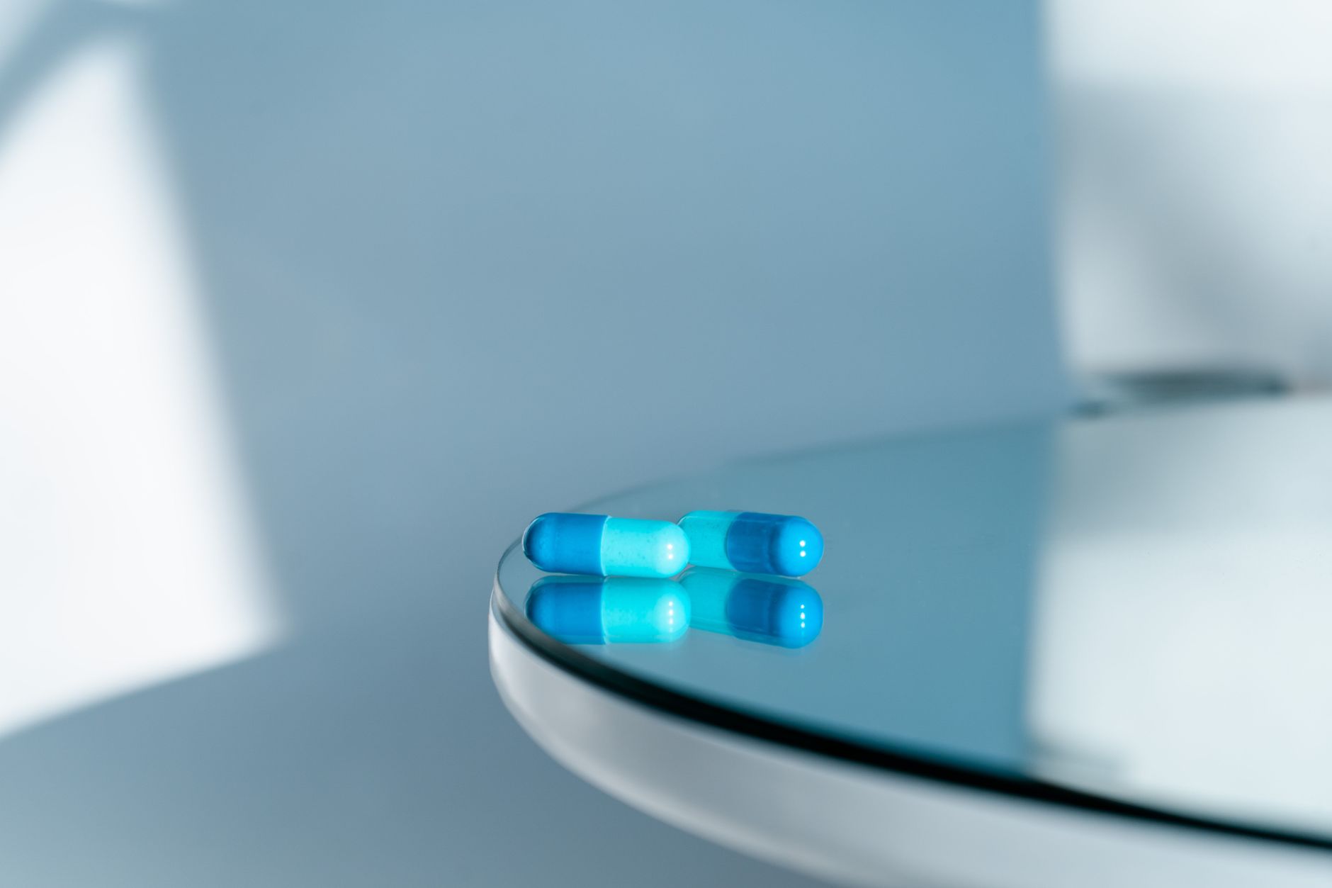 Close-up of two blue capsules on a shiny round glass table, reflecting light.
