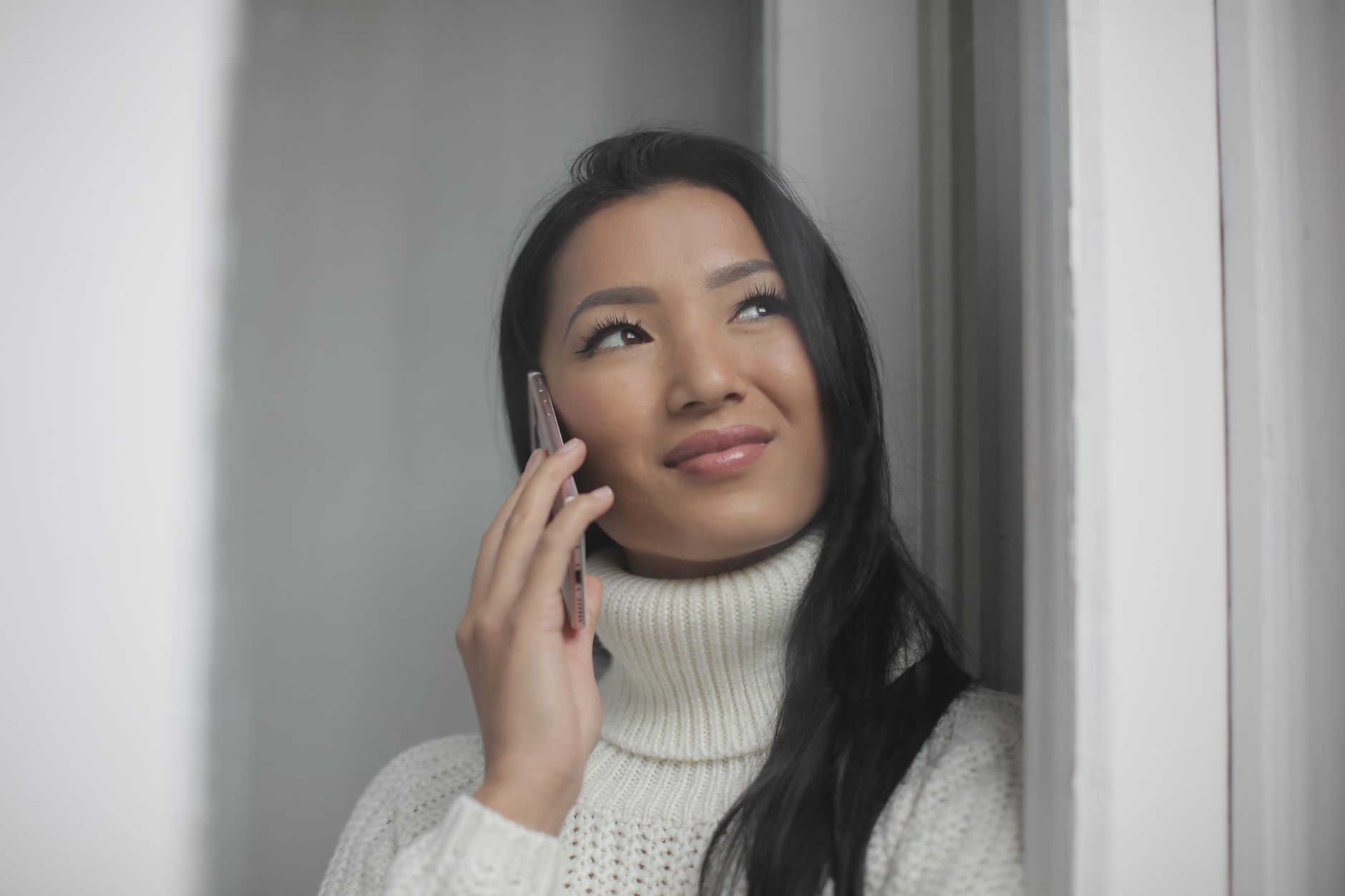 Asian woman in a white knit turtleneck smiling while talking on her phone by a window.