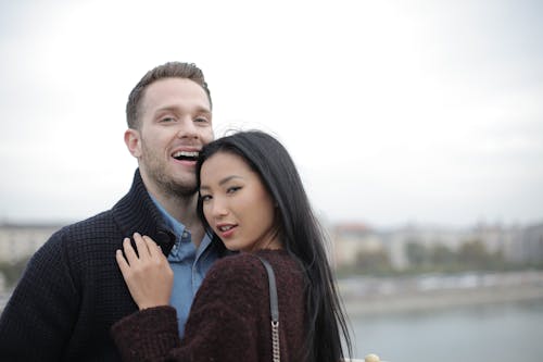 Positive young multiracial couple in casual warm clothes standing close to each other on street and looking at camera