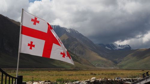 Free Georgian flag waving in Kvemo Okrokana with stunning mountainous backdrop. Stock Photo