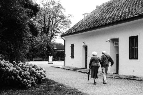 Free A black and white photograph of an elderly couple walking on a path outside a white building. Stock Photo