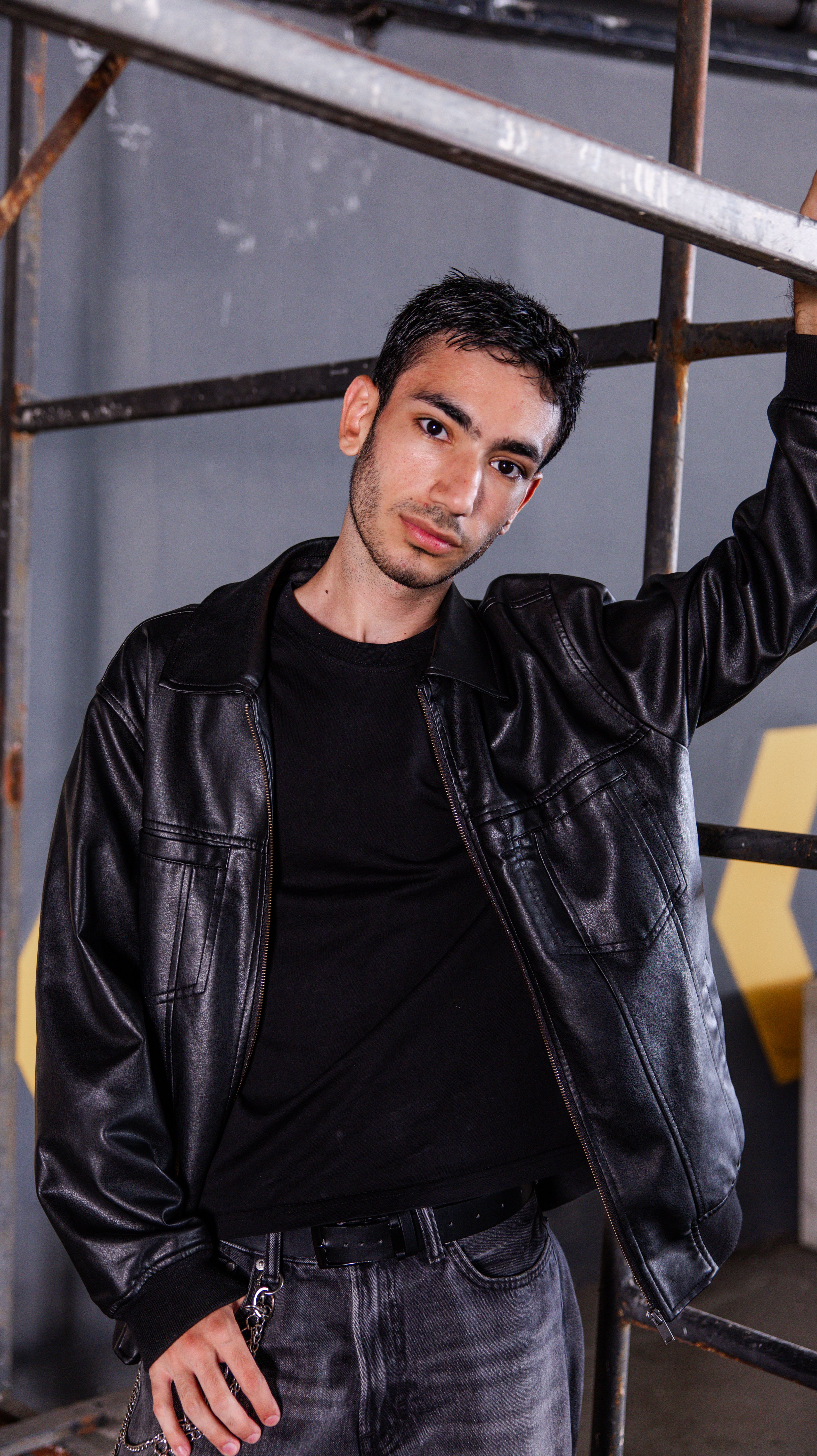 A fashionable male model posing casually in an industrial setting with a leather jacket.