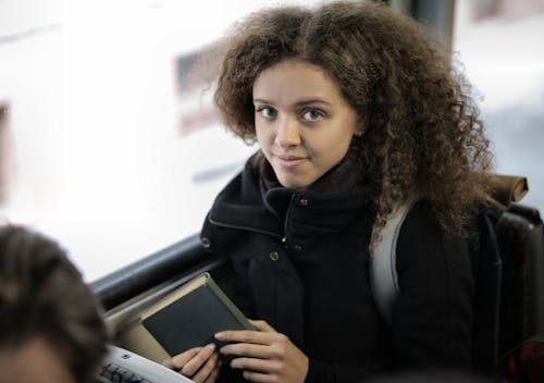 From above view of positive young lady with curly hair in casual warm coat sitting with book on passenger seat near window in bus and looking at camera