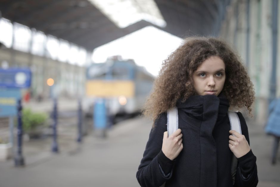 Teenager with curly hair wearing a trench coat stands at a train station, looking thoughtful.