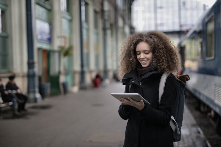 Cheerful Young Lady Browsing Tablet While Waiting For Train On Platform