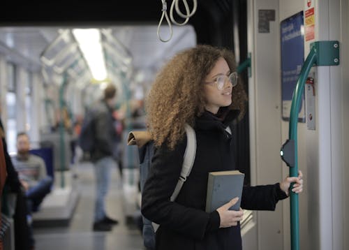 Positive young lady with curly hair in casual warm coat and eyeglasses riding train with backpack and book during travel