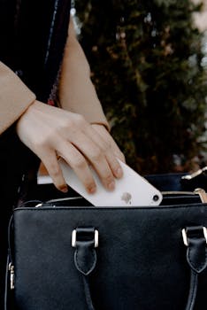 A hand carefully places a smartphone into a black leather handbag outdoors.