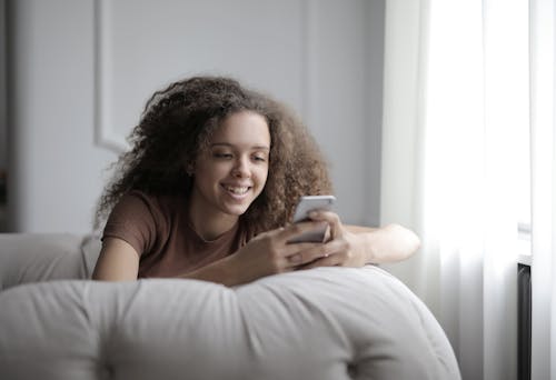 Free Curly teenage girl in casual clothes smiling while browsing mobile phone resting on cozy sofa in living room at home Stock Photo