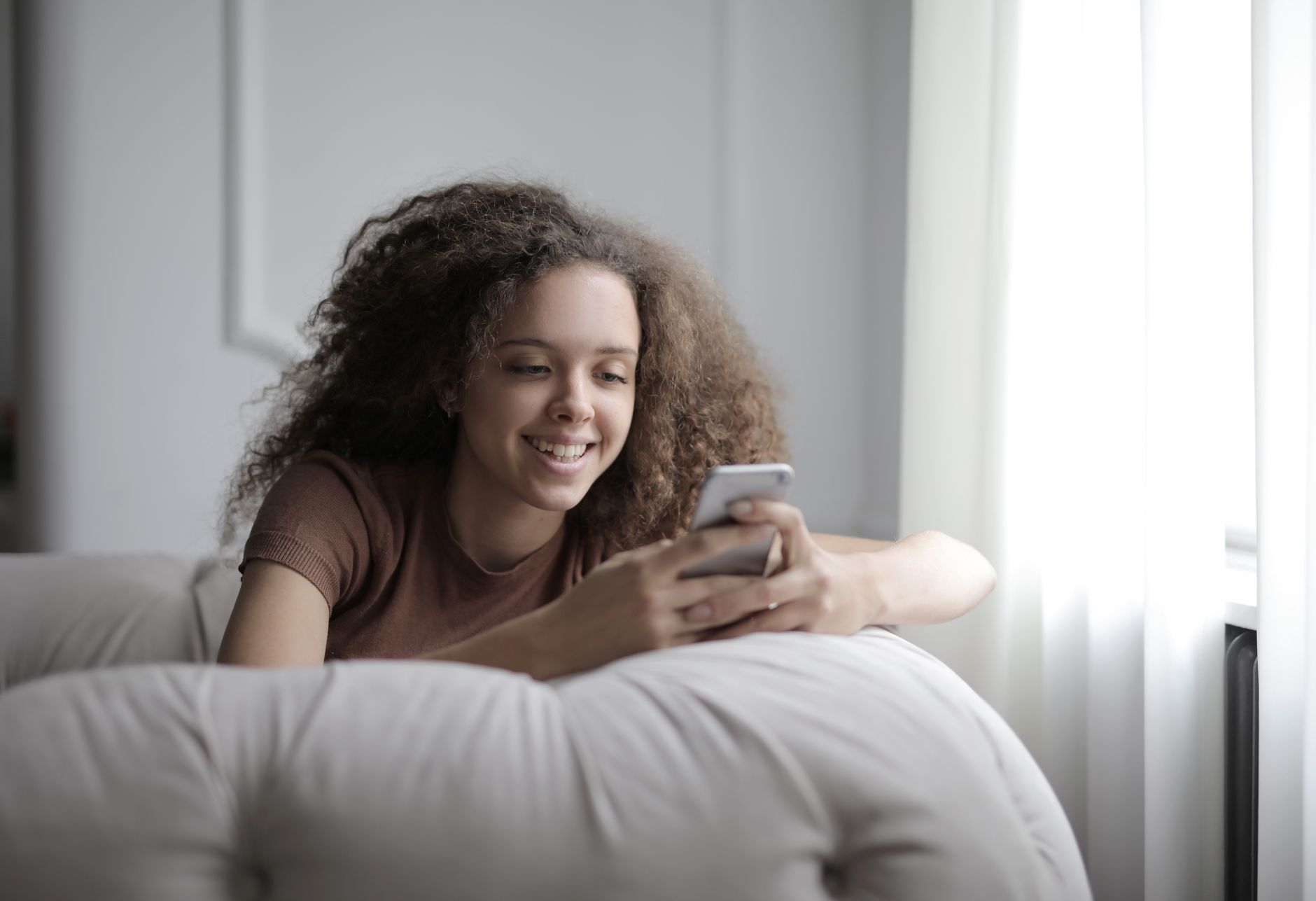 Smiling young woman lying on a sofa, looking at her smartphone screen near a window