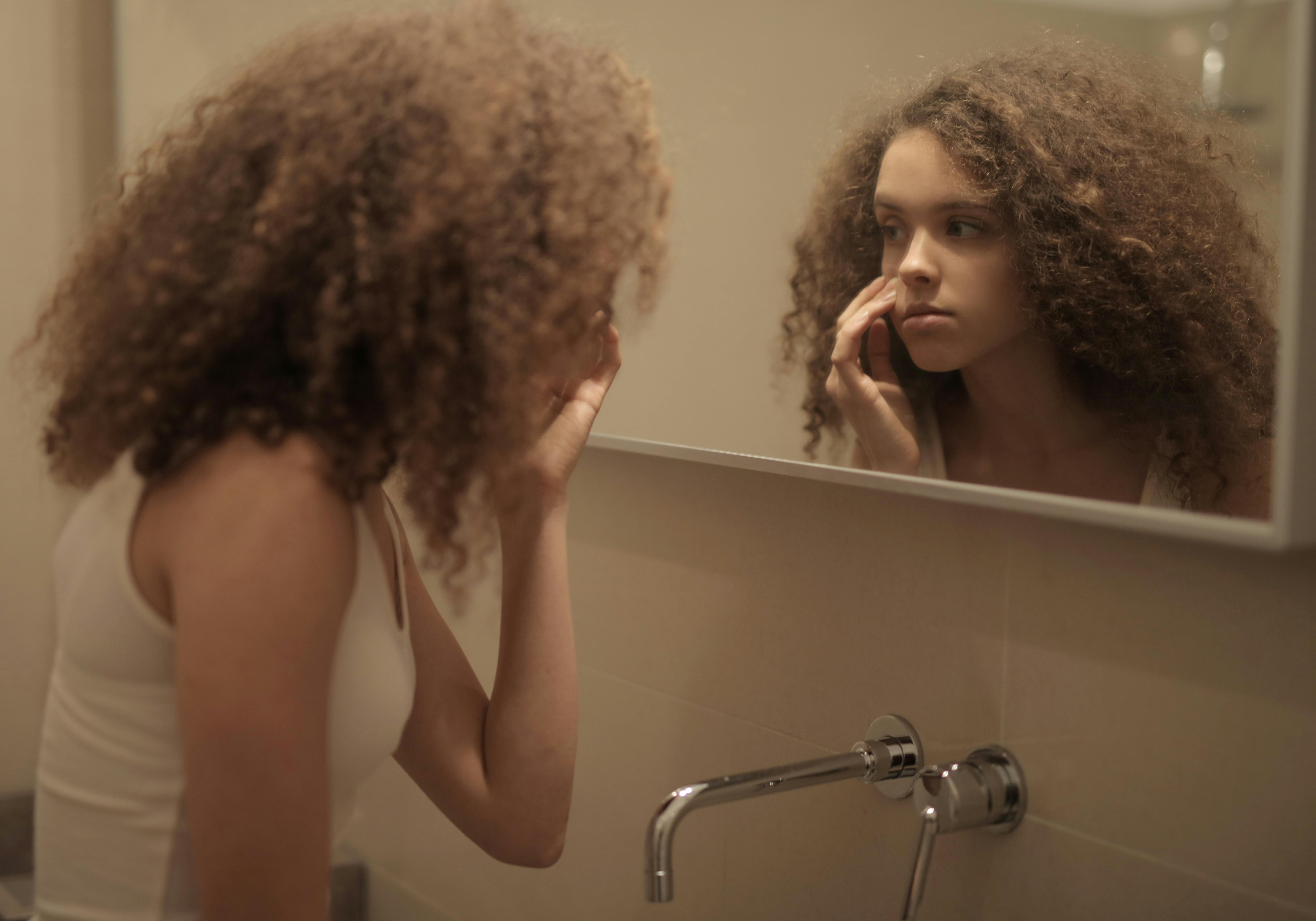 Teenage girl with curly hair examines her face in the bathroom mirror, reflecting contemplation.