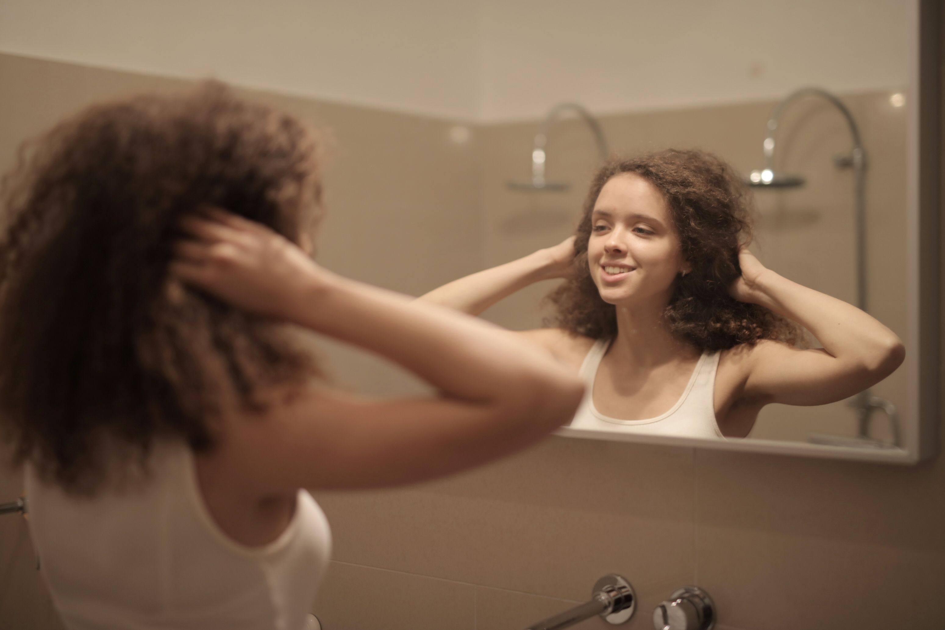 A woman with curly hair smiles at her reflection in the bathroom mirror, enjoying a relaxing moment.