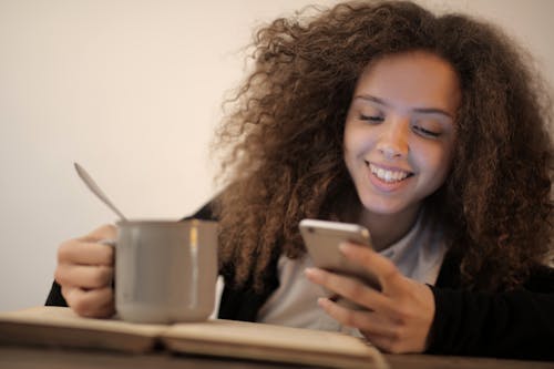 Free Cheerful young woman using mobile phone while drinking cacao at home Stock Photo