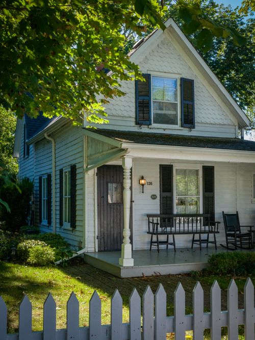 Free A picturesque white house with a picket fence surrounded by greenery and sunlight. Stock Photo