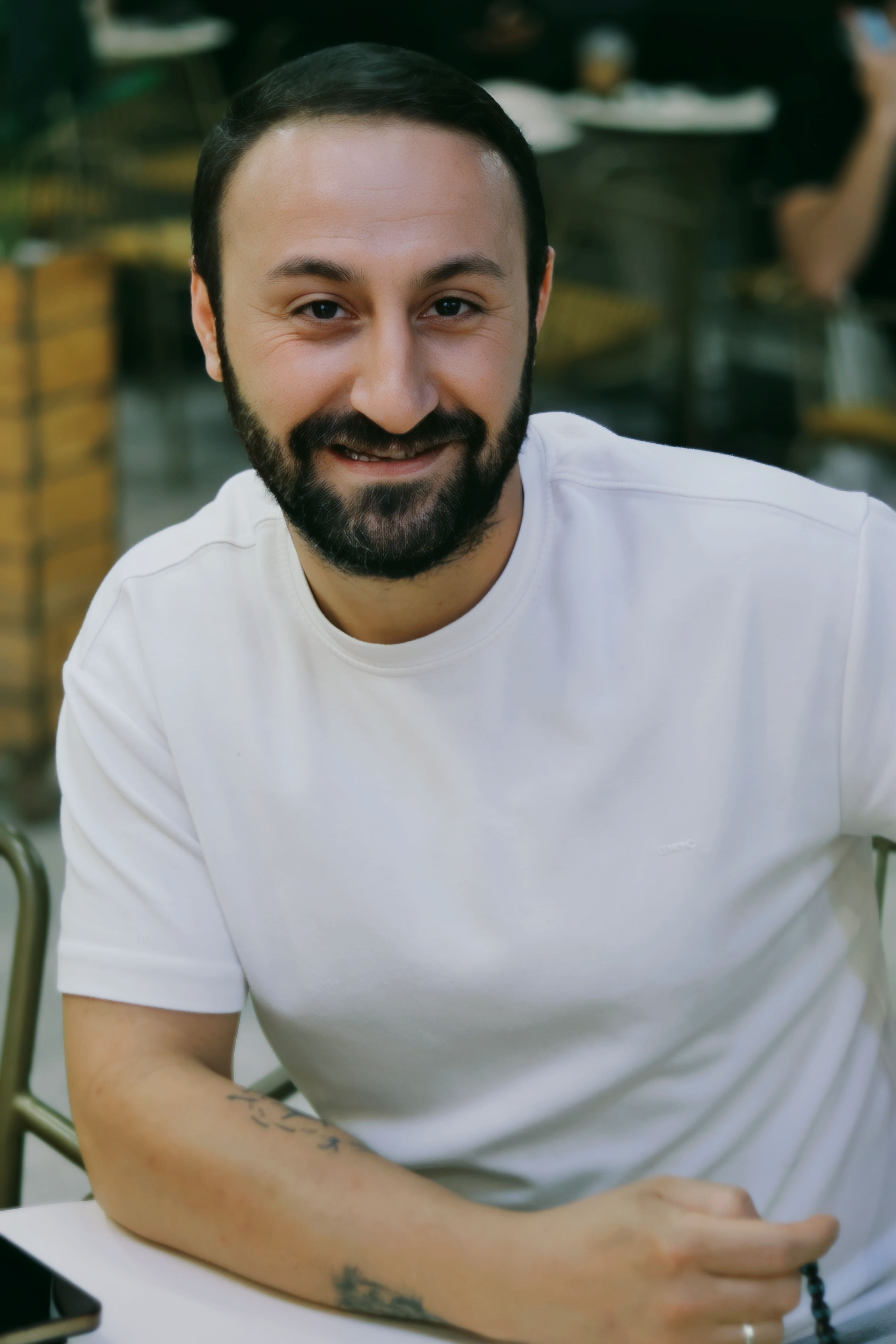 A casually dressed man seated and smiling indoors at a cafe setting.