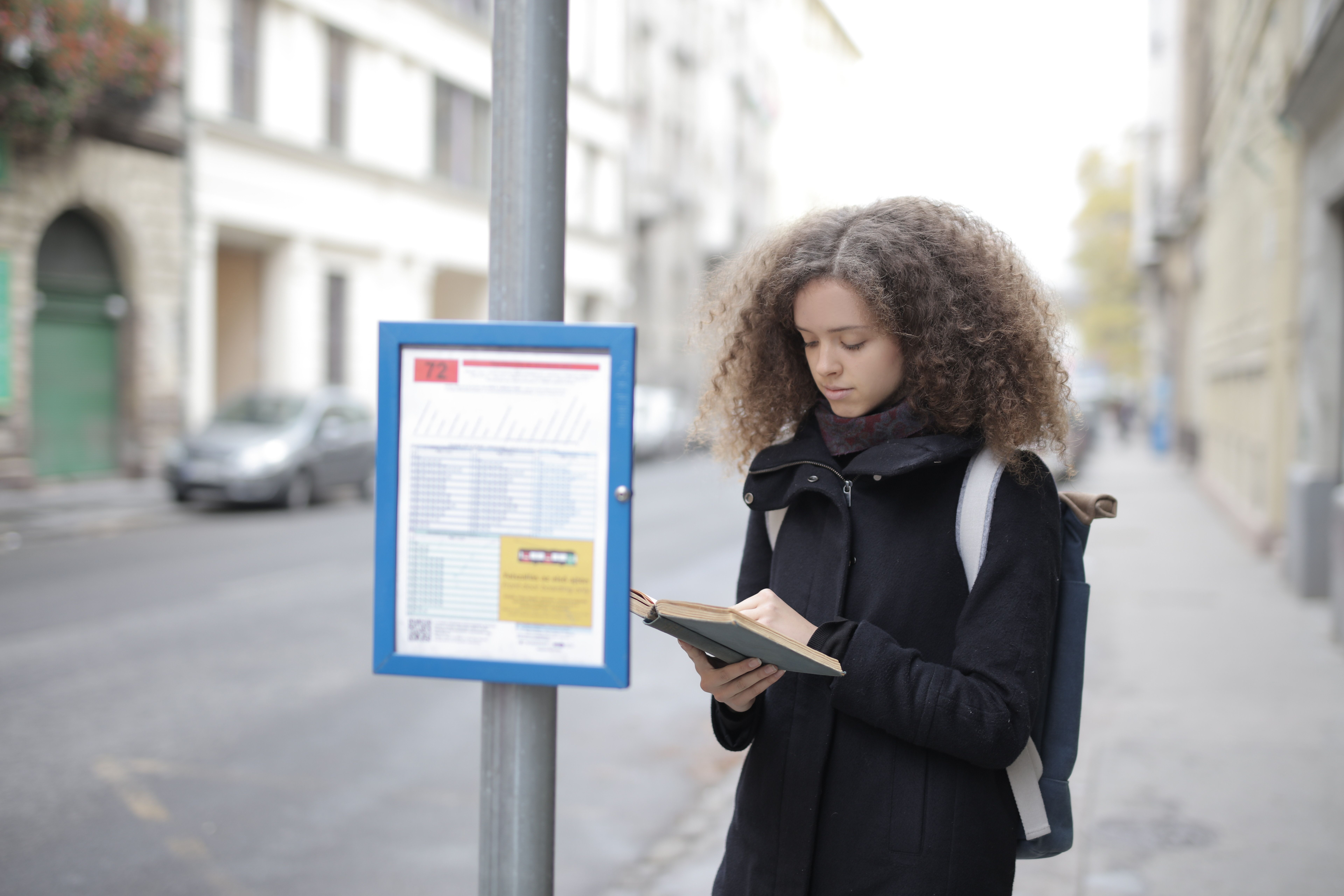A young woman with curly hair reads a book while standing at a bus stop on a city street.