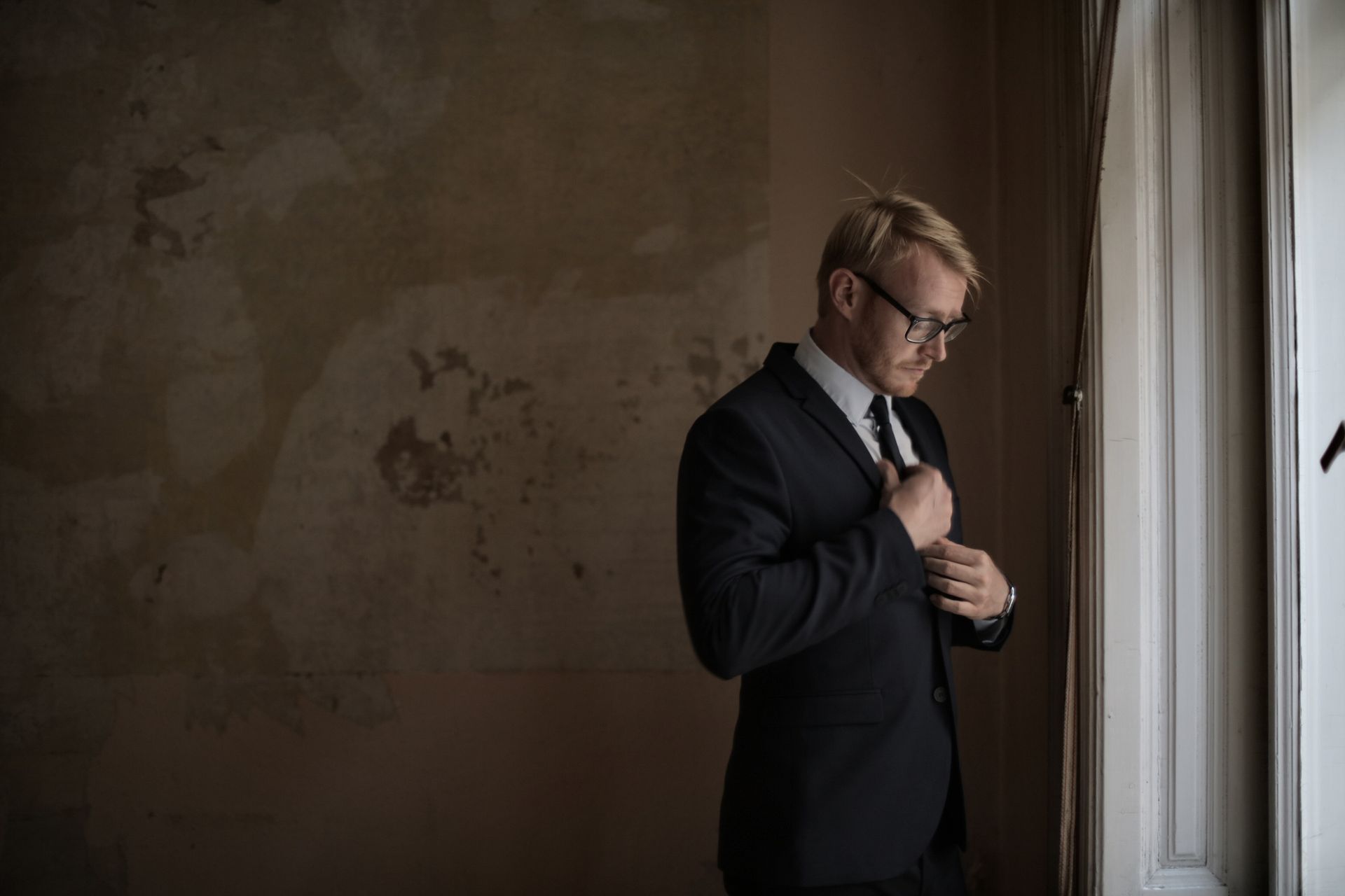 Side view of male realtor in formal wear bowing head and straightening tie while standing near windowsill in old building