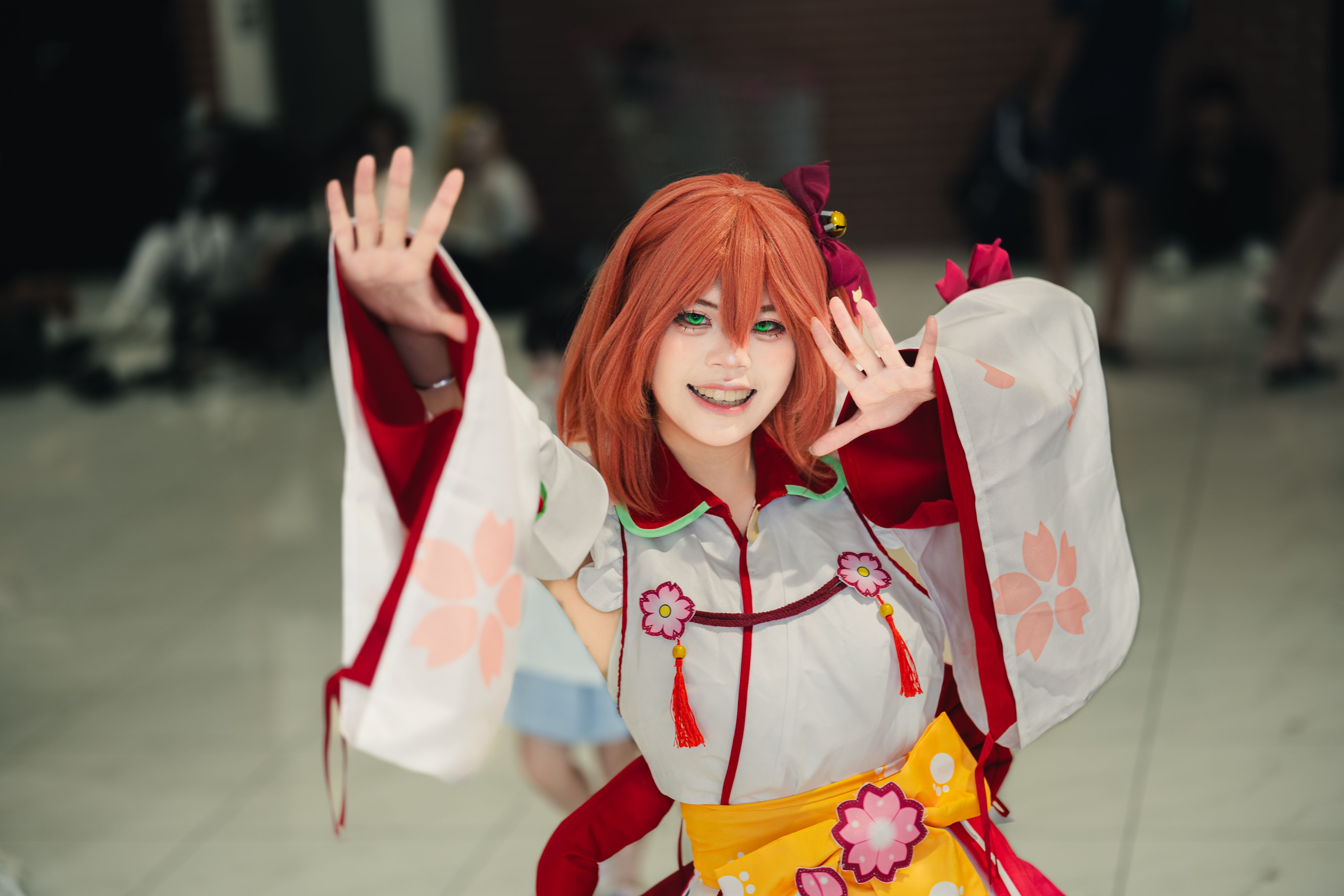 A smiling cosplayer in vibrant costume poses energetically at an anime convention indoors.