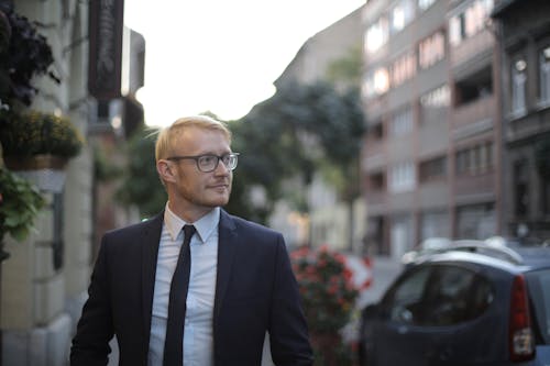 Free Positive young man in formal suit and eyewear looking away while strolling along sidewalk near car and historical buildings in city Stock Photo