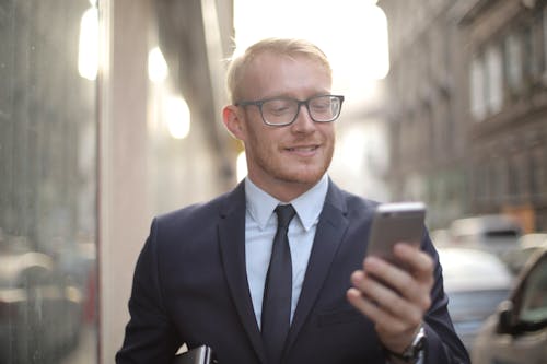 Free Smiling male entrepreneur in eyeglasses browsing smartphone in downtown Stock Photo