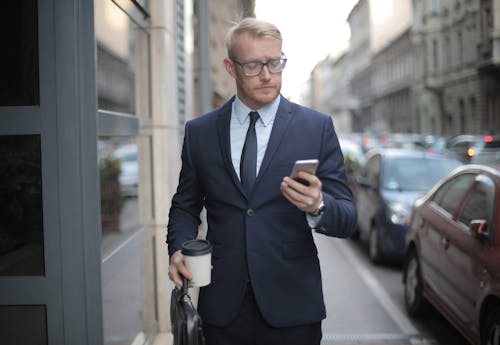 Serious businessman using cellphone walking takeaway coffee in city street