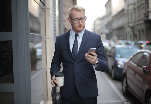 Free Focused young man in classy suit and glasses standing near modern building and browsing smartphone while having coffee break in downtown Stock Photo
