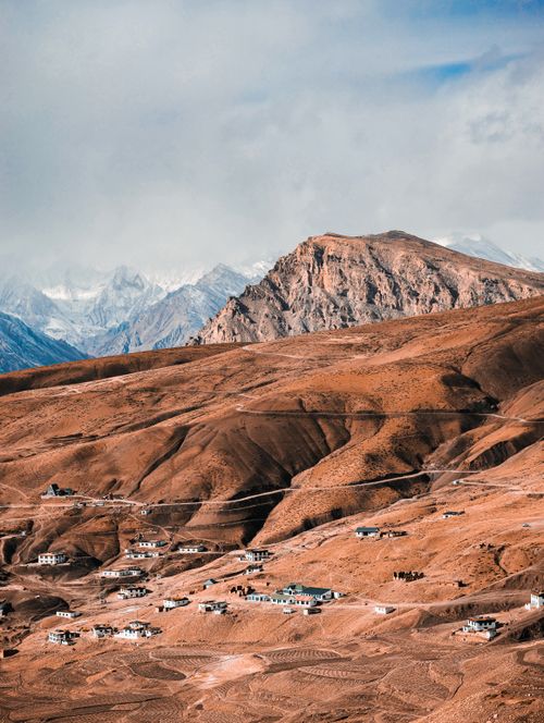 Free Aerial view of remote village nestled in rugged Spiti Valley mountains with snow-capped peaks in the background. Stock Photo