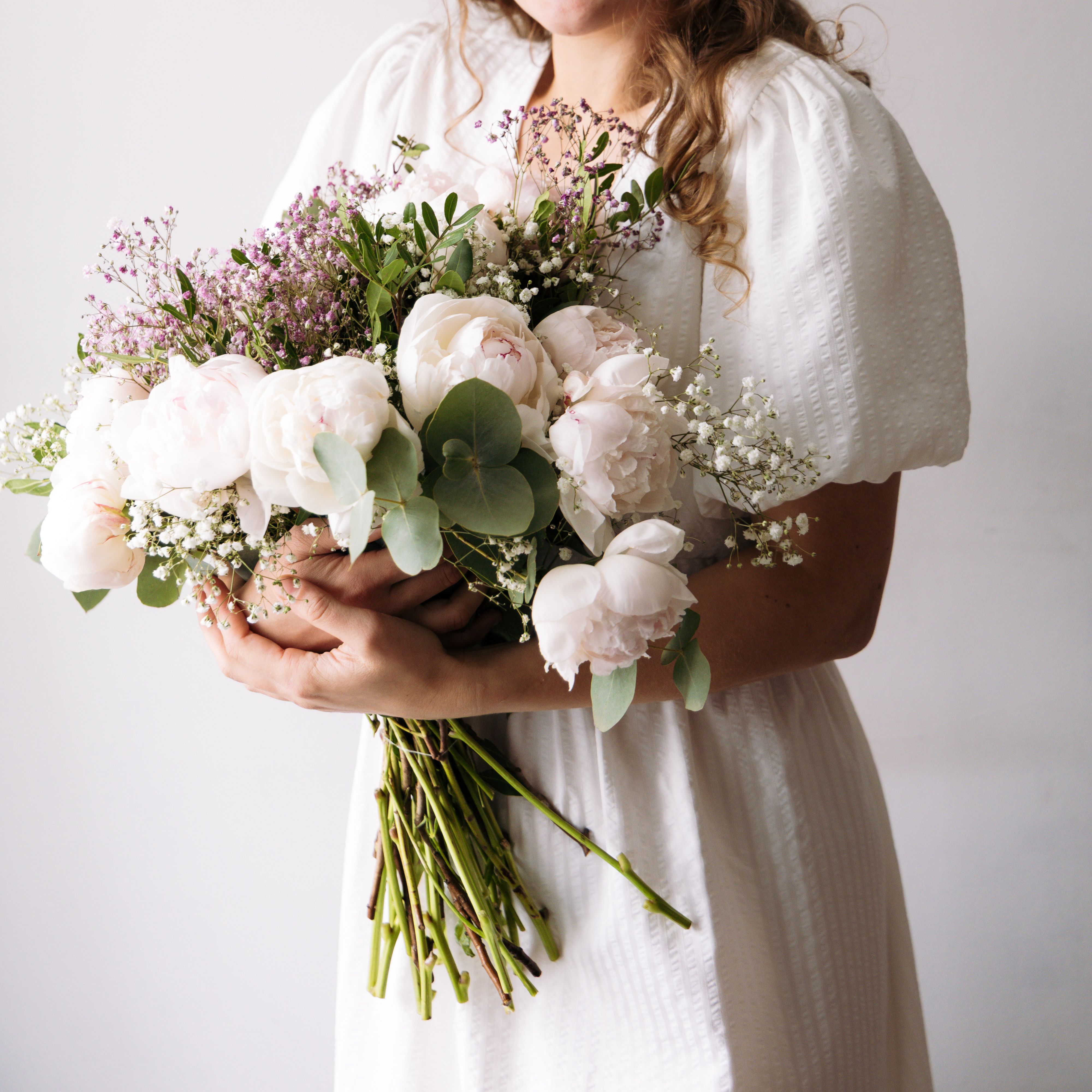 A woman in a white dress holds a bouquet of peonies and eucalyptus, symbolizing elegance and purity.