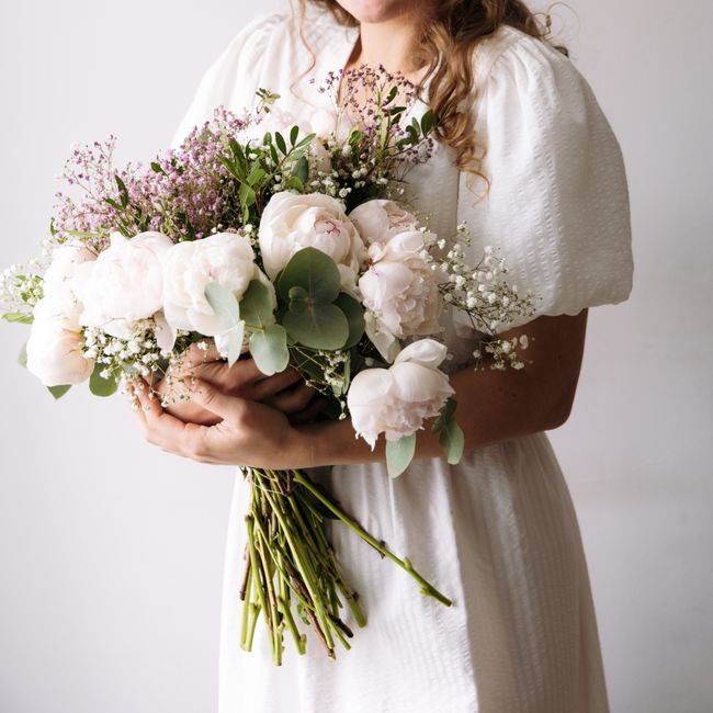 A woman in a white dress holds a bouquet of peonies and eucalyptus, symbolizing elegance and purity.