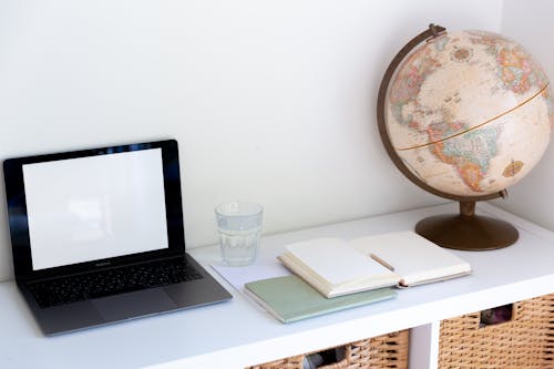 Laptop near glass of water opened blank notebook and retro globe on white cabinet