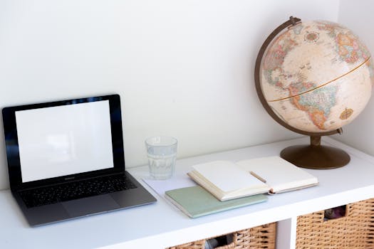 Clean and organized workspace with a laptop, globe, notebooks, and glass of water on a white desk.