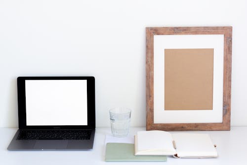 Laptop with blank screen and glass of water placed on white desk near empty frame and blank notepad against white wall
