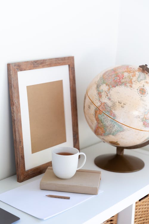 Free Vintage globe near blank frame and cup of tea placed on book in office Stock Photo