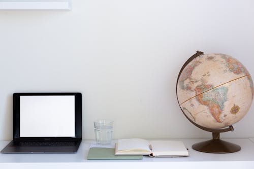 Creative composition of computer with empty screen placed near notebook with white papers and globe with glass of water in between
